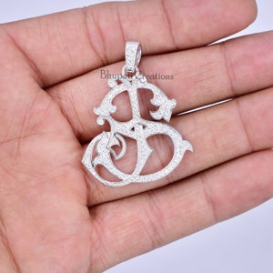 May include: A silver pendant with a decorative design, set with small, sparkling stones. The pendant has an elaborate, flowing shape with the text "Bhupati Creations" visible. The pendant is held in a hand.