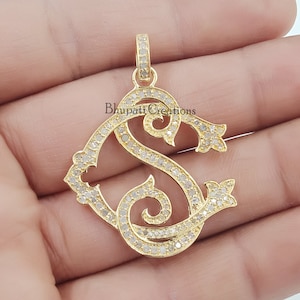 Peut inclure: Un pendentif en or avec une lettre 'S' sertie de diamants dans une police d'écriture décorative. Le pendentif est placé sur un fond uni.