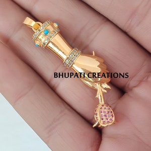 Op de afbeelding: Gouden hanger met een hand die een roos vasthoudt. De hand is versierd met turquoise en heldere stenen. De roos is gemaakt van roze stenen. De hanger is gelabeld met "BHUPATI CREATIONS".