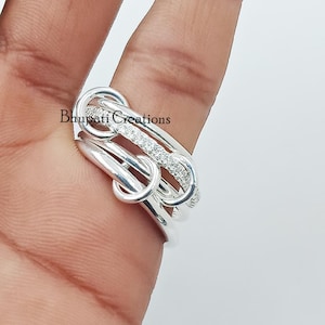 Puede incluir: Un anillo de plata con un diseño único de eslabones, con una fila de pequeñas piedras transparentes. El anillo se lleva en un dedo, con el nombre de la marca "Bhupati Creations" visible. El diseño del anillo es moderno y llamativo.