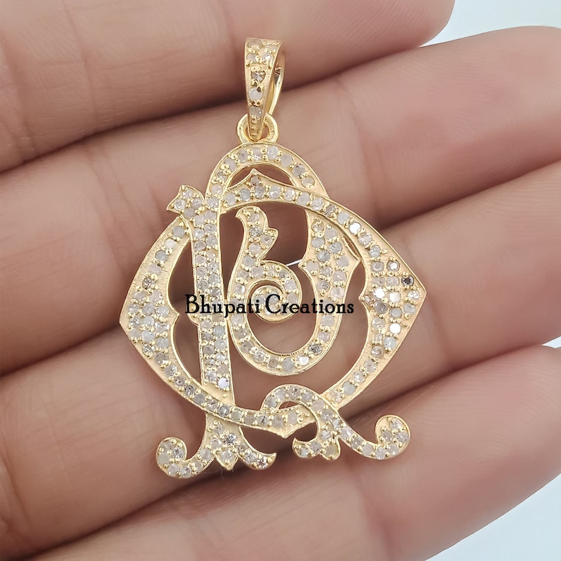 May include: A gold pendant featuring a diamond-encrusted design of the letters "R" and "B" intertwined. The pendant is set against a white background. "Bhupati Creations" is printed in black text at the bottom of the image.