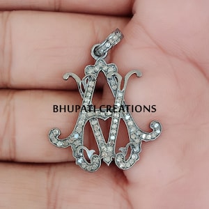 May include: A silver pendant with a monogram design featuring the letters "A" and "M" set with small diamonds. The pendant is on a hand.