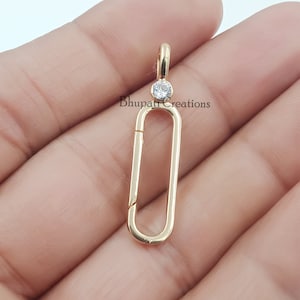 May include: A gold-coloured pendant with a rectangular shape and a small, round, clear gemstone at the top. The pendant has a clasp and a loop for attaching to a chain. The text "Bhupat Creations" is visible.