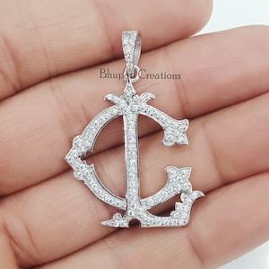 May include: Silver pendant featuring a large, ornate letter 'C' with a smaller 'L' inside. The pendant is covered in small, clear cubic zirconia stones.
