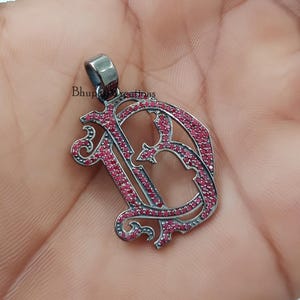 May include: Silver pendant with a large letter D made of silver and set with small red gemstones. The pendant is on a hand.