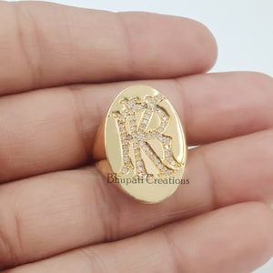 Op de afbeelding: Een gouden ovale ring met de letters "KR" bezet met kleine diamanten. De ring is op een hand.