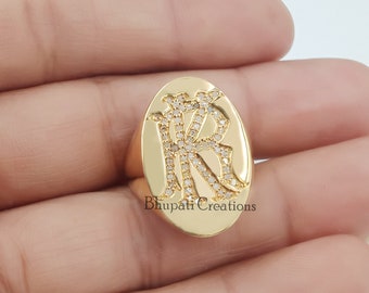 May include: A gold oval ring with the letters "KR" set with small diamonds. The ring is on a hand.