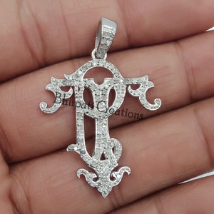 May include: A silver pendant with a diamond-encrusted design featuring the letters "PT". The pendant is set against a blurred background of a hand.