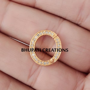 May include: A gold-colored, circular jewelry clasp with a ring of small, clear stones. The clasp is open, revealing the locking mechanism. The text "BHUPATI CREATIONS" is printed in black across the bottom of the image.