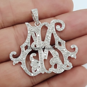 May include: A silver pendant with a monogram design featuring the letters "M" and "A" set with small diamonds. The pendant is on a white background.