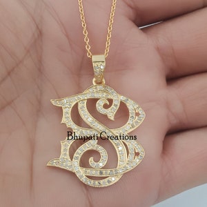 May include: A gold pendant necklace featuring a diamond-encrusted initial "S" charm. The pendant is in a cursive script style with a delicate design. The necklace is on a gold chain.