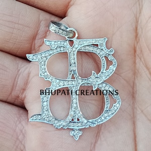 Puede incluir: Un colgante de plata con un diseño de monograma doble 'B', engastado con pequeños diamantes. El colgante está intrincadamente detallado y tiene una anilla para sujetarlo a una cadena.