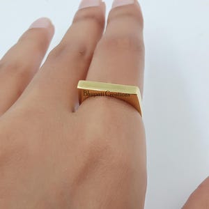 May include: A gold-coloured rectangular ring with the text "Bhupati Creations" engraved on the side. The ring is worn on a finger. The ring has a modern, minimalist design.
