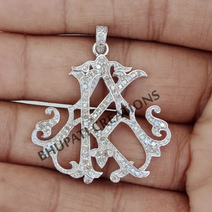 May include: A silver pendant with a diamond-encrusted monogram of the letters "AK". The pendant is in a classic, ornate style.