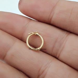 May include: A small, circular gold-toned metal ring featuring a hinged clasp mechanism at the top. The surface of the ring is smooth and polished, reflecting light. The text Bhupati Creations is printed in small black letters on the metal surface.