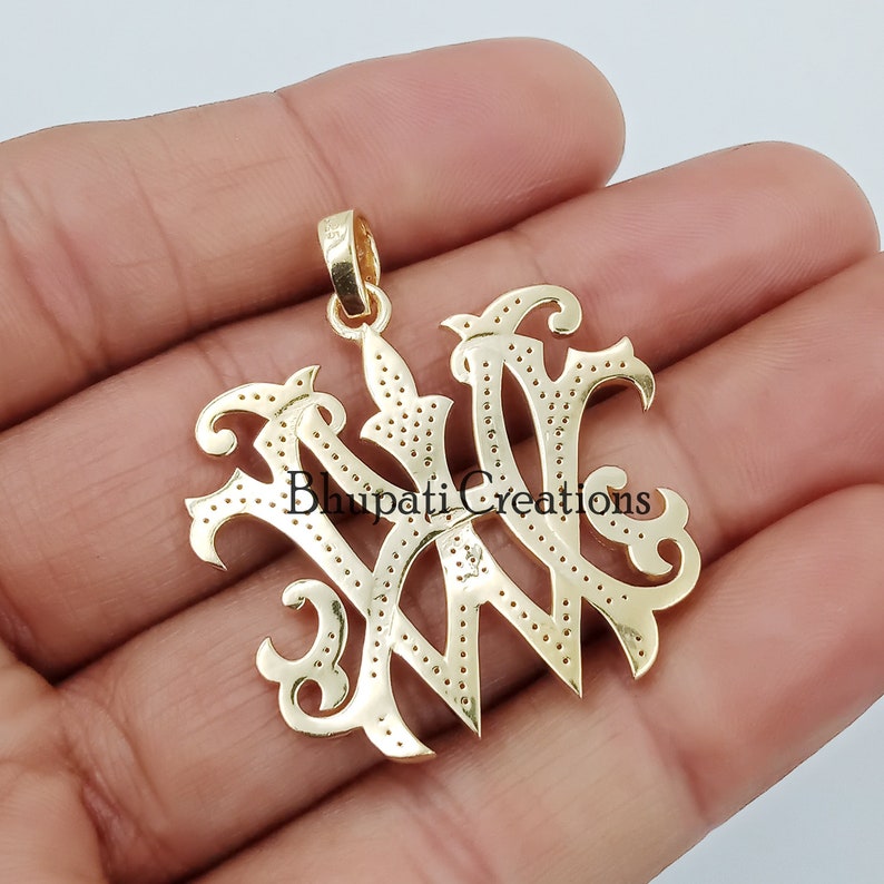 May include: Gold-toned pendant featuring a stylised monogram of the letters "WW" with a decorative design. The pendant has a bail for attaching to a chain.