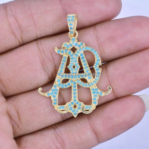 May include: Gold-toned pendant with the letters "AB" encrusted with small turquoise-colored stones. The pendant has a decorative design with a loop at the top for a chain. The text "BHUPATI CREATIONS" is visible.