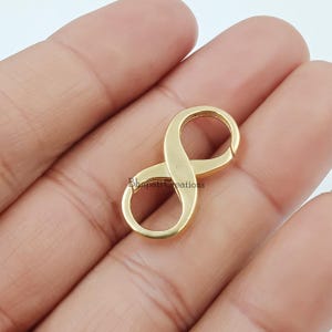 Op de afbeelding: Een goudkleurige oneindigheidssluiting, vastgehouden in een hand. De sluiting heeft de vorm van een oneindigheidssymbool, met een gladde, gepolijste afwerking. De sluiting wordt waarschijnlijk gebruikt voor het maken van sieraden of als decoratief element.