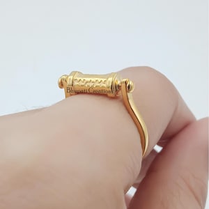 May include: A gold ring with a cylindrical central design and a curved band. The ring has the text "Bhupati Creations" on the central part. The ring is displayed on a finger against a white backdrop.