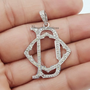 May include: A silver pendant with the intertwined letters "D" and "J" encrusted with small, sparkling diamonds. The pendant has a loop at the top for a chain and is held against a light background. The text "Bhupati Creations" is visible.