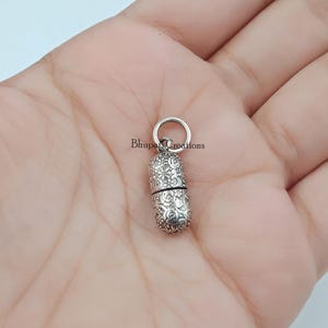 May include: A small, silver-coloured, cylindrical pill-shaped charm featuring an intricate, embossed floral pattern across its surface. The charm includes a circular jump ring at the top for attachment and displays the text Bhupati Creations.