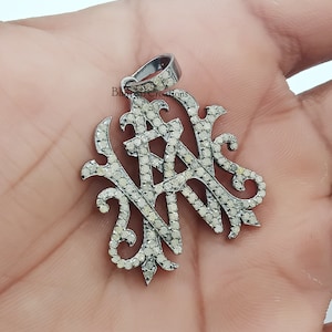 Può includere: Un ciondolo d'argento con un design a monogramma incastonato con piccoli diamanti bianchi.