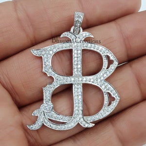 May include: A silver pendant in the shape of the letter "B" encrusted with small, sparkling stones. The pendant has an ornate design with a loop at the top for a chain. The text "Bhupat Creations" is visible on the pendant.