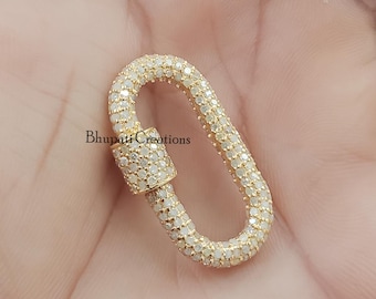Puede incluir: Un cierre ovalado dorado, cubierto de pequeñas piedras blancas brillantes. El cierre tiene una sección con bisagras para abrir y cerrar. El texto "Bhupati Creations" es visible en el cierre.