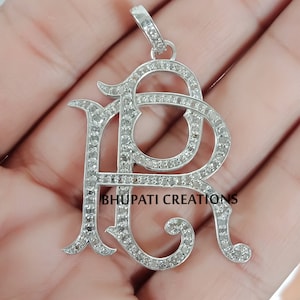 Peut inclure: Un pendentif en argent avec les lettres "PR" en écriture cursive, serti de petits diamants. Le pendentif est sur une chaîne et est tenu par une main. Le texte "BHUPATI CREATIONS" est visible sous le pendentif.
