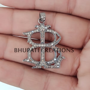 Peut inclure: Pendentif en argent avec un grand motif en forme de lettre B, incrusté de petits diamants. Le pendentif est sur une main.