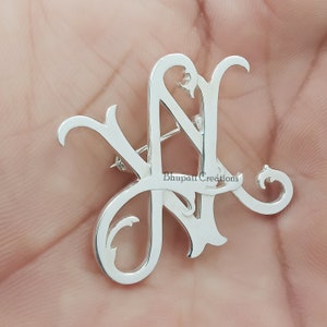 Peut inclure: Broche en argent avec monogramme représentant les lettres "N" et "L" entrelacées dans une police d'écriture décorative. La broche est en argent et se ferme à l'aide d'une épingle. Le texte "Bhupati Creations" est visible sur la broche.