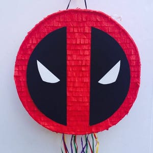 Pode incluir: Uma pinhata redonda vermelha com o design da máscara do Deadpool. A máscara é composta por um oval preto com duas formas de olhos brancos angulares. A pinhata é decorada com papel crepom vermelho e tem serpentinas coloridas.
