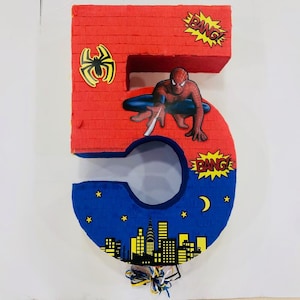 Puede incluir: Una piñata de cartón roja y azul con forma de número 5. La piñata está decorada con un personaje de Spiderman, un paisaje urbano y la palabra "Bang!" en bocadillos de diálogo amarillos.