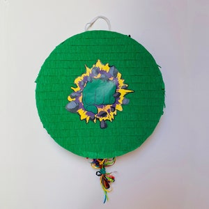 May include: Green and yellow pinata with a cartoon image of a green fist breaking through a wall. The pinata is round and has a fringed edge.
