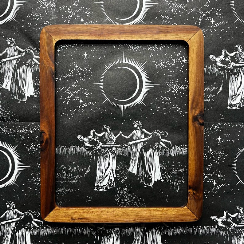 A framed print featuring a black and white illustration of female figures in white gowns dancing in a circle under a crescent moon and starry sky. The print is set in a rectangular wooden frame with rounded corners.