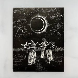 Black and white art print depicting a celestial scene. A crescent moon with radiating lines is centered above 5 female figures in flowing robes, holding hands in a circle. The background is a starry night sky, with a field of tall grass in the foreground.