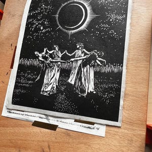Black and white art print depicting a celestial scene. A crescent moon with radiating lines is centered above 5 female figures in flowing robes, holding hands in a circle. The background is a starry night sky, with a field of tall grass in the foreground.