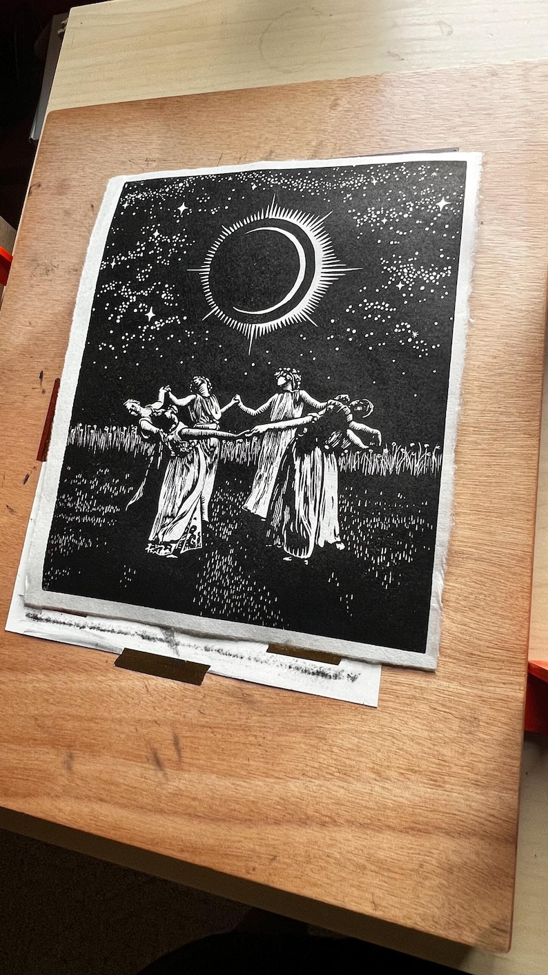 Black and white art print depicting a celestial scene. A crescent moon with radiating lines is centered above 5 female figures in flowing robes, holding hands in a circle. The background is a starry night sky, with a field of tall grass in the foreground.