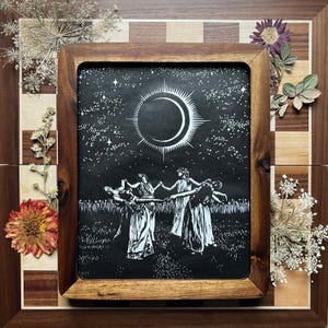 Framed image of a Black and white art print depicting a celestial scene. A crescent moon with radiating lines is centered above 5 female figures in flowing robes, holding hands in a circle. The background is a starry night sky, with a field of tall grass in the foreground.