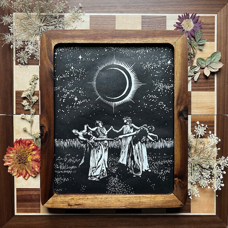 Framed image of a Black and white art print depicting a celestial scene. A crescent moon with radiating lines is centered above 5 female figures in flowing robes, holding hands in a circle. The background is a starry night sky, with a field of tall grass in the foreground.