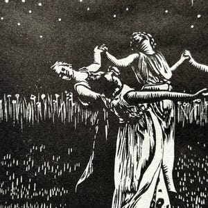 A detail shot of a Black and white art print depicting a celestial scene. A crescent moon with radiating lines is centered above 5 female figures in flowing robes, holding hands in a circle. The background is a starry night sky, with a field of tall grass in the foreground.
