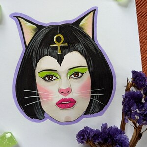May include: A hand-drawn illustration of a person with black hair and cat ears. The person is wearing green eyeshadow and pink blush. A gold ankh symbol is on their forehead.