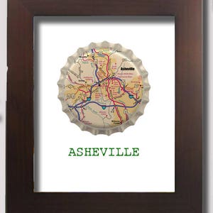Puede incluir: Una impresión enmarcada de un mapa de Asheville, Carolina del Norte, impreso en una tapa de botella. El texto "ASHEVILLE" está impreso debajo del mapa.
