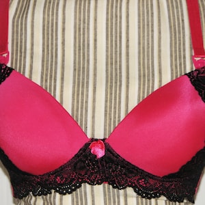 May include: A pink bra with black lace trim. The bra has adjustable straps and a hook and eye closure in the back.