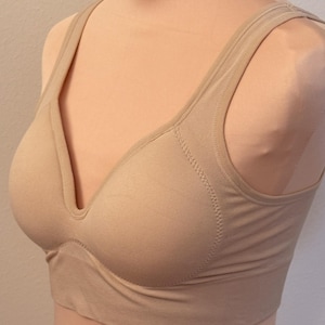 May include: A beige wireless bra featuring a deep V-neckline, wide shoulder straps, and a seamless construction. The garment has a solid tan color with reinforced stitching along the neckline and side panels, providing a smooth and supportive fit.