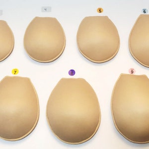 May include: A collection of eight beige bra inserts in various sizes, arranged in two rows. Each insert is oval-shaped with a rounded bottom and a slightly flattened top. Size labels 2, 4, 5, 6, 7, 8, and 9 are visible above or near each insert.