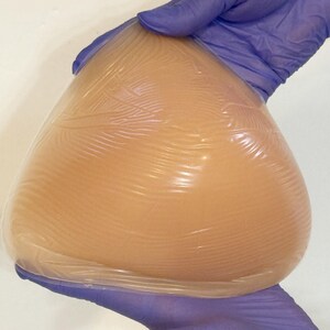 May include: A teardrop-shaped silicone breast form with a smooth, translucent, light beige surface. The material is flexible and soft, featuring a subtle, fine-lined texture across the front surface, designed for use as a prosthetic insert.