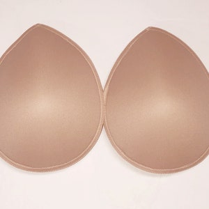 May include: Two beige, teardrop-shaped bra inserts. Each insert has a smooth, padded surface and is outlined with a stitched border. The inserts are designed to enhance the shape and size of the bust.