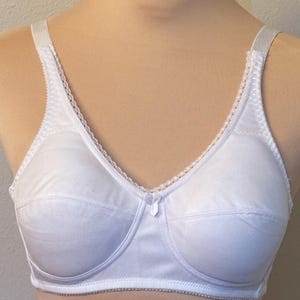 May include: A white brassiere featuring multi-part cups with horizontal seams, a delicate lace trim along the neckline, and a small central bow detail. The garment includes adjustable shoulder straps and a structured underband for support.