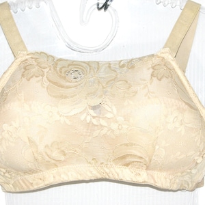 May include: A beige bra with floral lace overlay. The bra features adjustable straps and a full-coverage design. The lace pattern includes roses and other floral elements, creating a delicate and feminine look. The bra is displayed on a clear hanger.